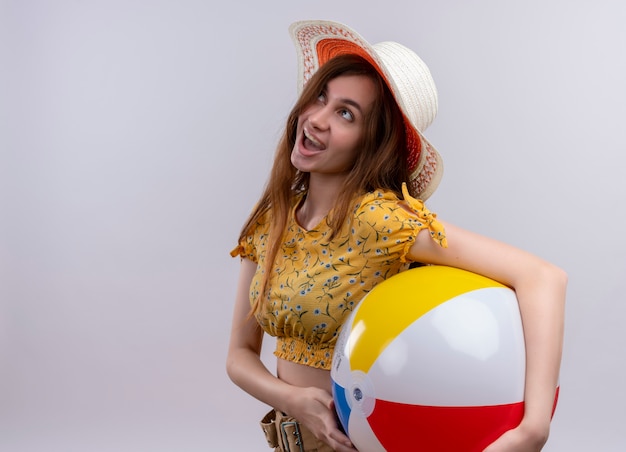 Chica joven emocionada con sombrero sosteniendo una pelota de playa mirando hacia arriba en un espacio en blanco aislado con espacio de copia