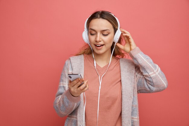 Chica joven emocionada usando y tocando auriculares sosteniendo y mirando el teléfono móvil