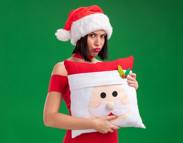 Chica joven enfurruñada con sombrero de santa sosteniendo la almohada de santa claus con los labios fruncidos aislados en la pared verde