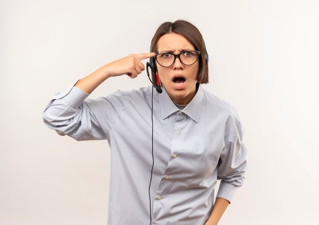 Chica joven enojada del centro de llamadas con gafas y auriculares poniendo el dedo en la sien aislado en blanco con espacio de copia