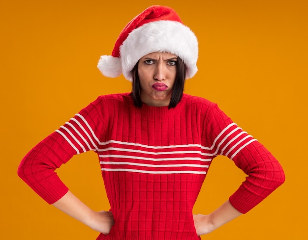 Chica joven enojada con gorro de Papá Noel manteniendo las manos en la cintura aislado en la pared naranja