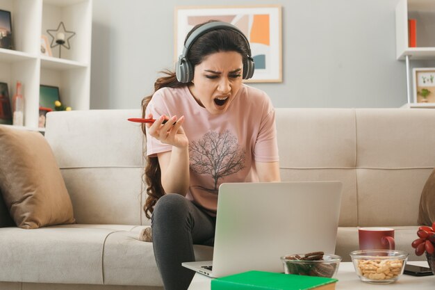Chica joven enojada usando audífonos con lápiz portátil usado sentado en el sofá detrás de la mesa de café en la sala de estar