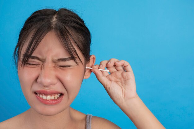 La chica joven es la oreja que escoge con la esponja de algodón en la pared azul, estudio.