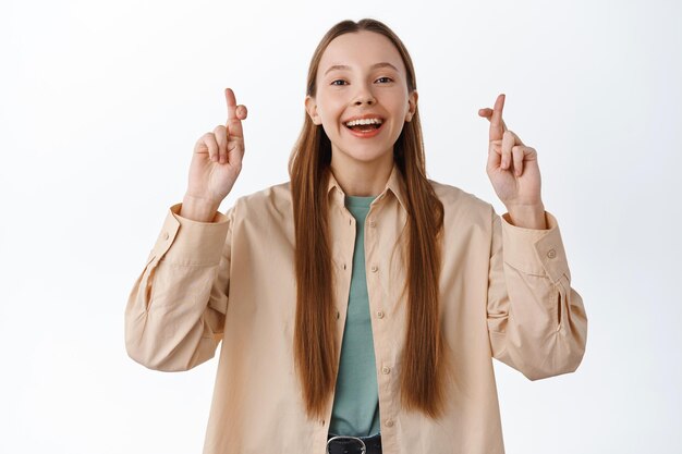 Chica joven esperanzada esperando los resultados del examen con los dedos cruzados, pidiendo deseos, pidiendo suerte, de pie optimista contra el fondo blanco con una sonrisa sincera