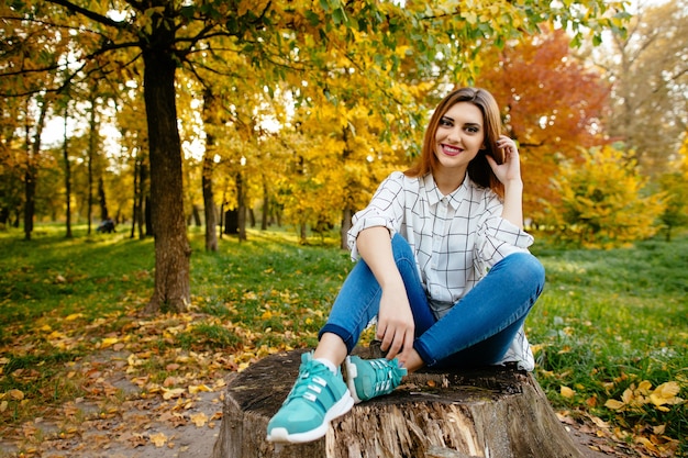 La chica joven se está sentando en un tocón en el parque del otoño.