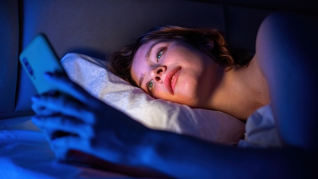 La chica joven está en su teléfono inteligente en la cama. Iluminación azul en la habitación.