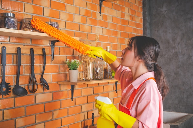 La chica joven está usando guantes amarillos mientras limpia la sala de cocina con plumero en su casa