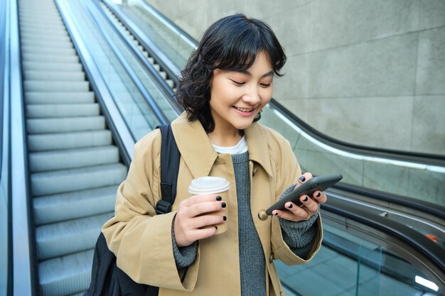 Chica joven con estilo con una taza de café bebe capuchino para ir baja las escaleras mecánicas y mira mobil