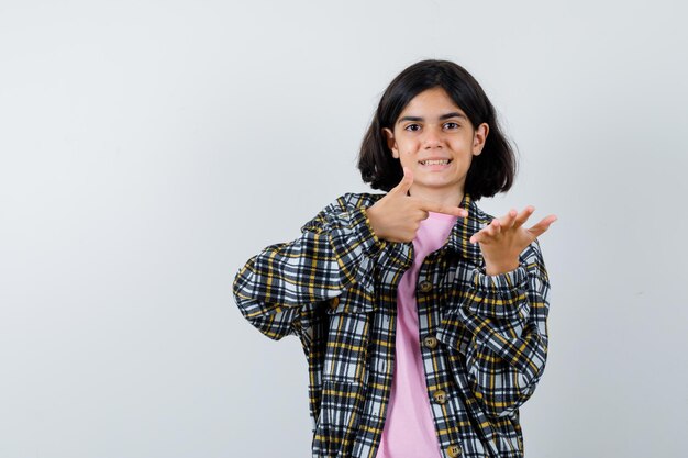 Chica joven estirando una mano como sosteniendo algo y apuntando con el dedo índice en camisa a cuadros y camiseta rosa y luciendo bonita, vista frontal.