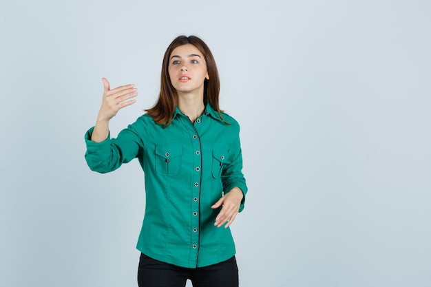 Chica joven estirando la mano como sosteniendo algo imaginario en blusa verde, pantalón negro y mirando enfocado, vista frontal.