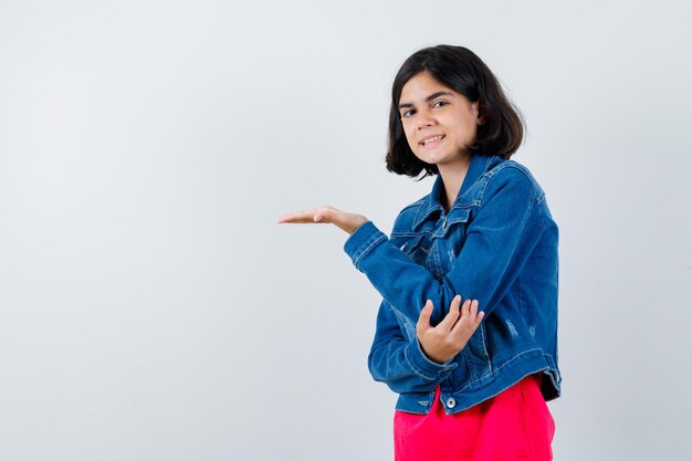 Chica joven estirando una mano como sosteniendo algo y sosteniendo la mano en el codo en camiseta roja y chaqueta de jean y mirando feliz, vista frontal.