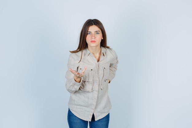 Chica joven estirando la mano de manera interrogante en camisa beige, jeans y mirando perplejo, vista frontal.