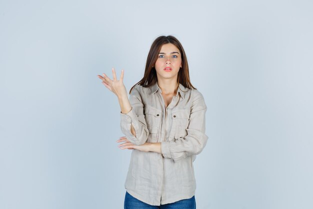 Chica joven estirando la mano de manera interrogante en camisa beige, jeans y mirando perplejo, vista frontal.
