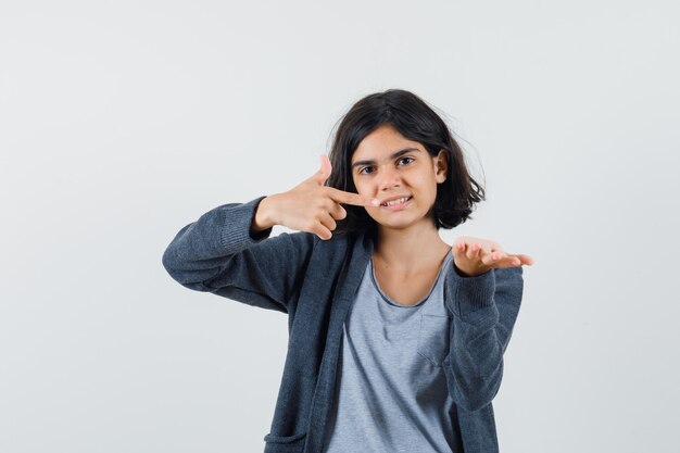 Chica joven estirando una mano mientras sostiene algo y apuntando hacia él con el dedo índice en una camiseta gris claro y una sudadera con capucha gris oscuro con cremallera y se ve linda