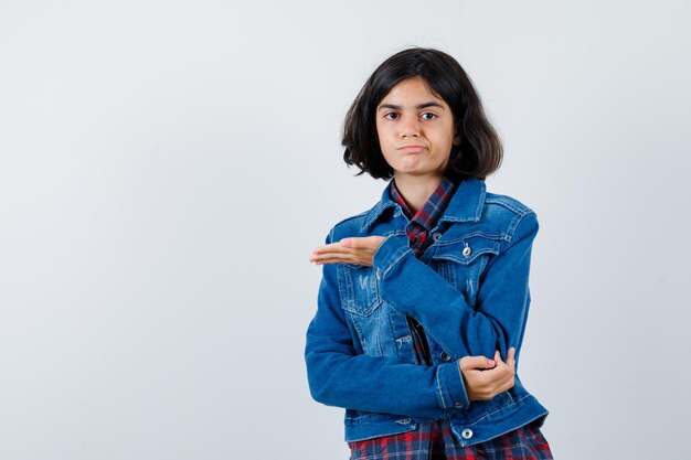 Chica joven estirando una mano mientras sostiene algo en camisa a cuadros y chaqueta de mezclilla y parece serio. vista frontal.