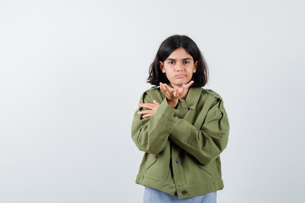 Chica joven estirando la mano hacia en suéter gris, chaqueta de color caqui, pantalón de mezclilla y mirando serio, vista frontal.