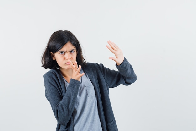 Chica joven estirando las manos como parando algo en una camiseta gris claro y una sudadera con capucha frontal con cremallera gris oscuro y mirando asustado