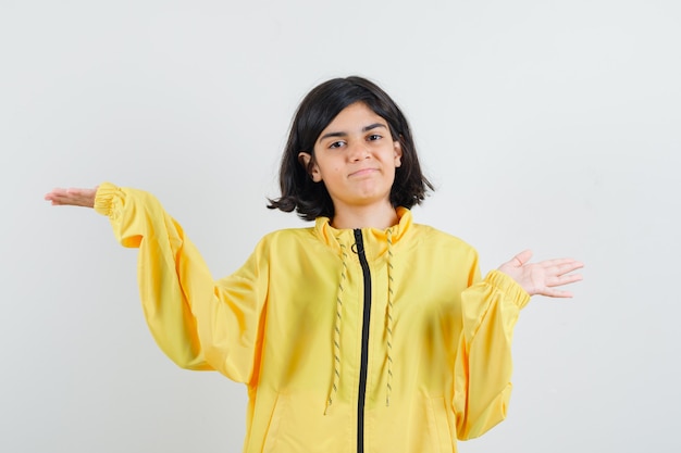 Chica joven estirando las manos como sosteniendo algo imaginario en chaqueta de bombardero amarilla y mirando serio.