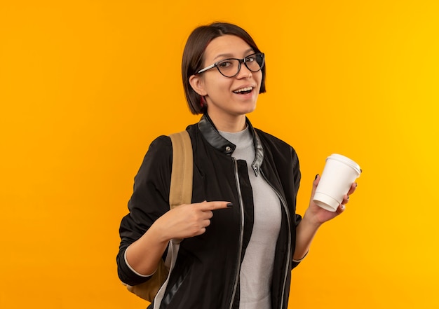 Chica joven estudiante alegre con gafas y bolsa trasera sosteniendo y apuntando a una taza de café de plástico aislada en naranja