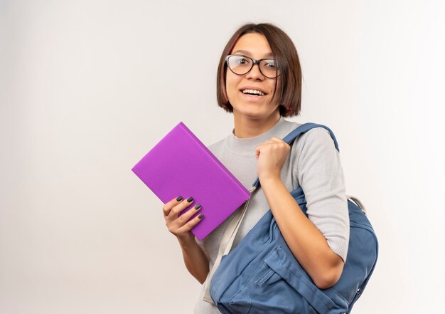 Chica joven estudiante alegre con gafas y bolsa trasera sosteniendo libro aislado en blanco