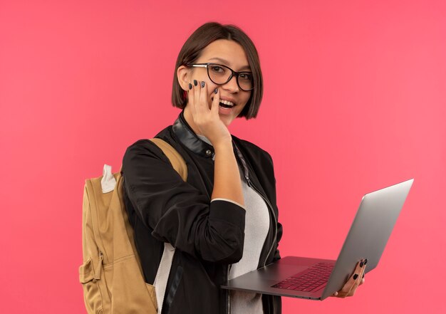 Chica joven estudiante alegre con gafas y bolsa trasera sosteniendo portátil poniendo la mano en la mejilla aislada en rosa