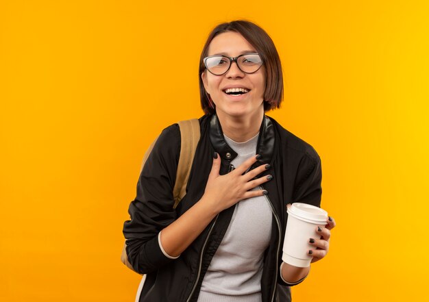 Chica joven estudiante alegre con gafas y bolsa trasera sosteniendo una taza de café de plástico poniendo la mano en el pecho aislado en naranja