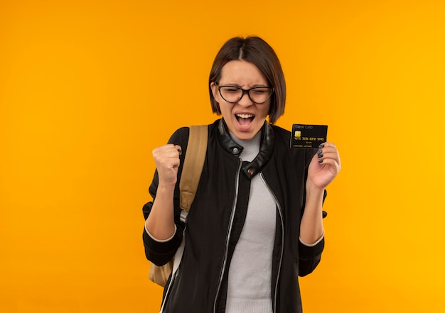 Chica joven estudiante alegre con gafas y bolsa trasera con tarjeta de crédito apretando el puño con los ojos cerrados aislados en naranja
