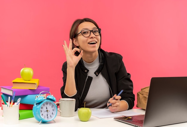 Chica joven estudiante alegre con gafas sentado en el escritorio haciendo bien signo aislado en rosa