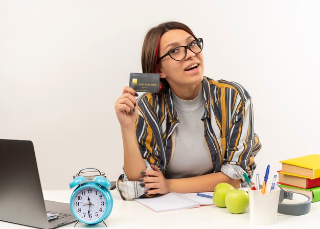 Chica joven estudiante alegre con gafas sentado en el escritorio mostrando tarjeta de crédito aislado en blanco