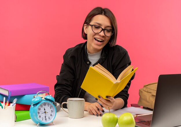 Chica joven estudiante alegre con gafas sentado en el escritorio sosteniendo y leyendo el libro haciendo los deberes aislados en rosa