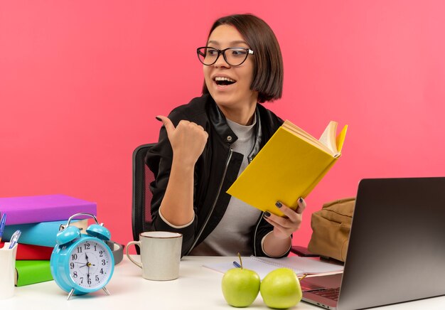 Chica joven estudiante alegre con gafas sentado en el escritorio sosteniendo el libro haciendo los deberes mirando y apuntando al lado aislado en rosa