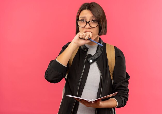 Chica joven estudiante ansiosa con gafas y bolsa trasera con bloc de notas y bolígrafo manteniendo la mano cerca de la boca aislada en rosa con espacio de copia