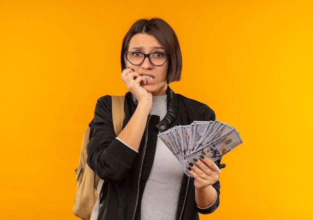 Chica joven estudiante ansiosa con gafas y bolsa trasera sosteniendo dinero poniendo la mano en la barbilla aislada en naranja con espacio de copia