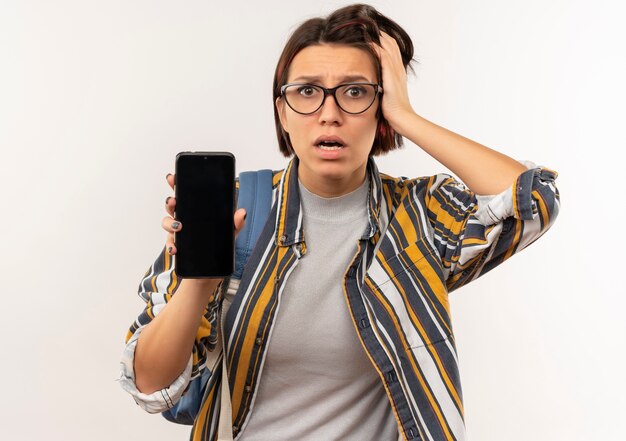 Chica joven estudiante ansiosa con gafas y bolso trasero mostrando teléfono móvil poniendo la mano en la cabeza aislada en blanco