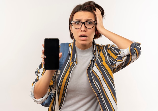 Chica joven estudiante ansiosa con gafas y bolso trasero mostrando teléfono móvil poniendo la mano en la cabeza aislada en blanco