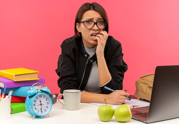 Chica joven estudiante ansiosa con gafas sentado en el escritorio haciendo los deberes poniendo la mano en los labios sosteniendo la pluma aislada en rosa