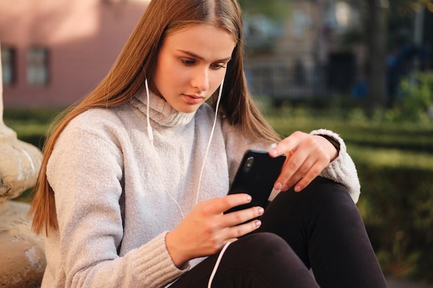 Chica joven estudiante atractiva en suéter acogedor usando cuidadosamente el teléfono celular durante el descanso de estudio al aire libre