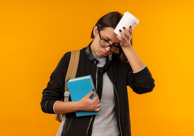 Chica joven estudiante cansada con gafas y bolsa trasera sosteniendo libro tocando la cabeza con la taza de café con los ojos cerrados aislado sobre fondo naranja