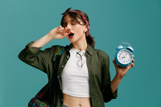 Chica joven estudiante cansada con pañuelo y mochila mostrando despertador poniéndose gafas en la blusa manteniendo la mano delante de la boca con los ojos cerrados bostezando aislada en el fondo azul