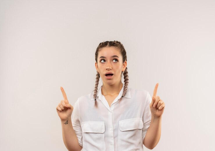 Foto gratuito chica joven estudiante con coletas en camisa blanca apuntando con el dedo índice hacia arriba mirando sorprendido de pie sobre la pared blanca