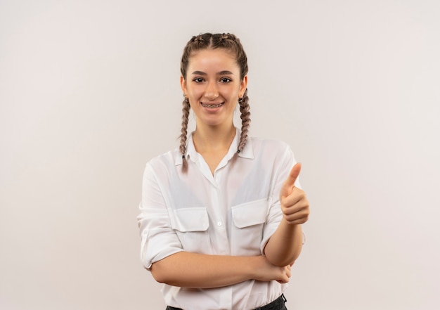 Chica joven estudiante con coletas en camisa blanca mirando al frente sonriendo mostrando los pulgares para arriba de pie sobre la pared blanca