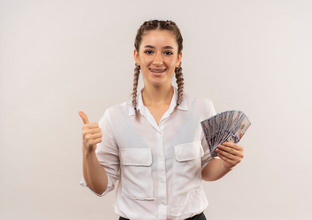 Foto gratuita chica joven estudiante con coletas en camisa blanca mostrando dinero en efectivo sonriendo mirando al frente mostrando los pulgares para arriba de pie sobre la pared blanca