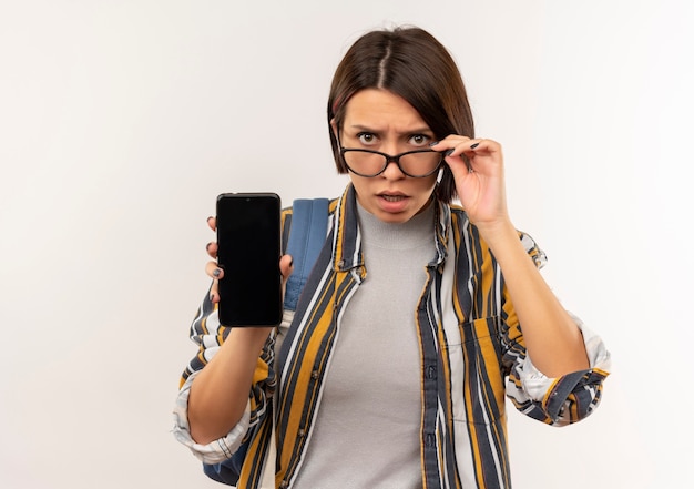 Chica joven estudiante confundida con gafas y bolso trasero mostrando teléfono móvil sosteniendo gafas aislado en blanco con espacio de copia