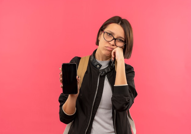 Chica joven estudiante disgustada con gafas y bolsa trasera sosteniendo teléfono móvil poniendo la mano en la mejilla aislada sobre fondo rosa con espacio de copia