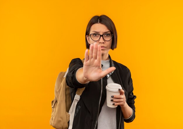 Chica joven estudiante disgustada con gafas y bolsa trasera con taza de café gesticulando parada en cámara aislada sobre fondo naranja con espacio de copia
