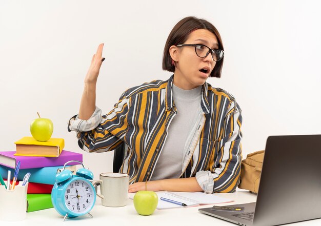 Chica joven estudiante disgustada con gafas sentado en un escritorio con herramientas universitarias gesticulando no y mirando al lado aislado sobre fondo blanco.