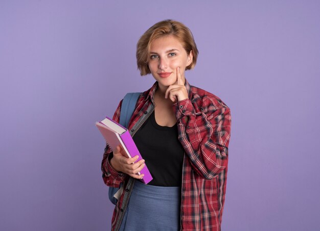 Chica joven estudiante eslava complacida con mochila pone el dedo en la cara sostiene el libro y el cuaderno