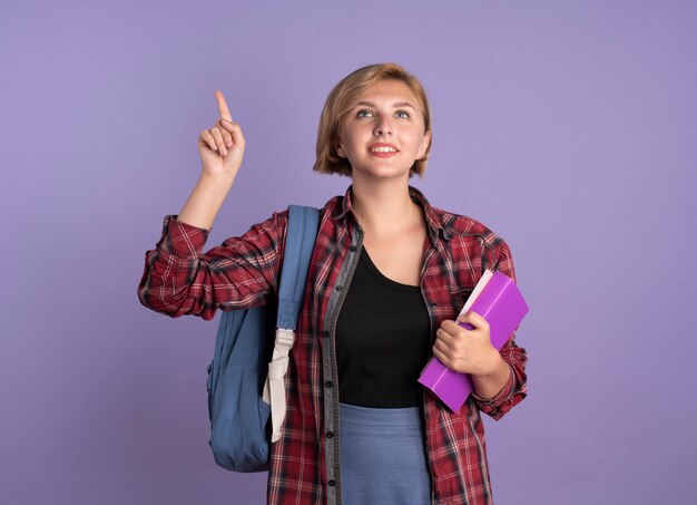 Chica joven estudiante eslava complacida con mochila sostiene el libro y el cuaderno apunta hacia arriba