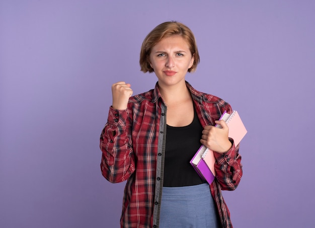 Foto gratuita chica joven estudiante eslava confiada mantiene el puño sostiene el libro y el cuaderno