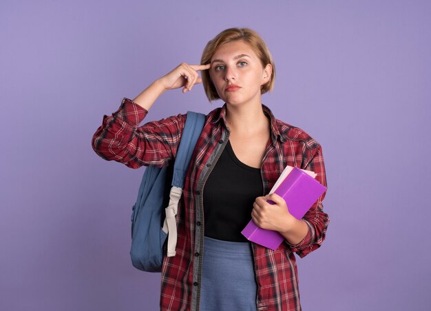 Chica joven estudiante eslava confiada que lleva la mochila pone el dedo en el templo sostiene el libro y el cuaderno